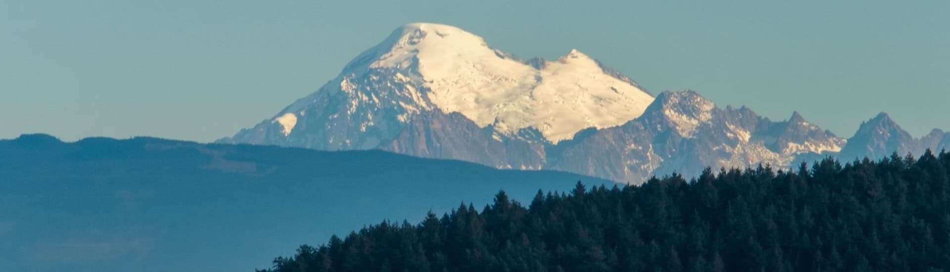 Snow-capped mountain peaks rise above a forested landscape under a clear sky.