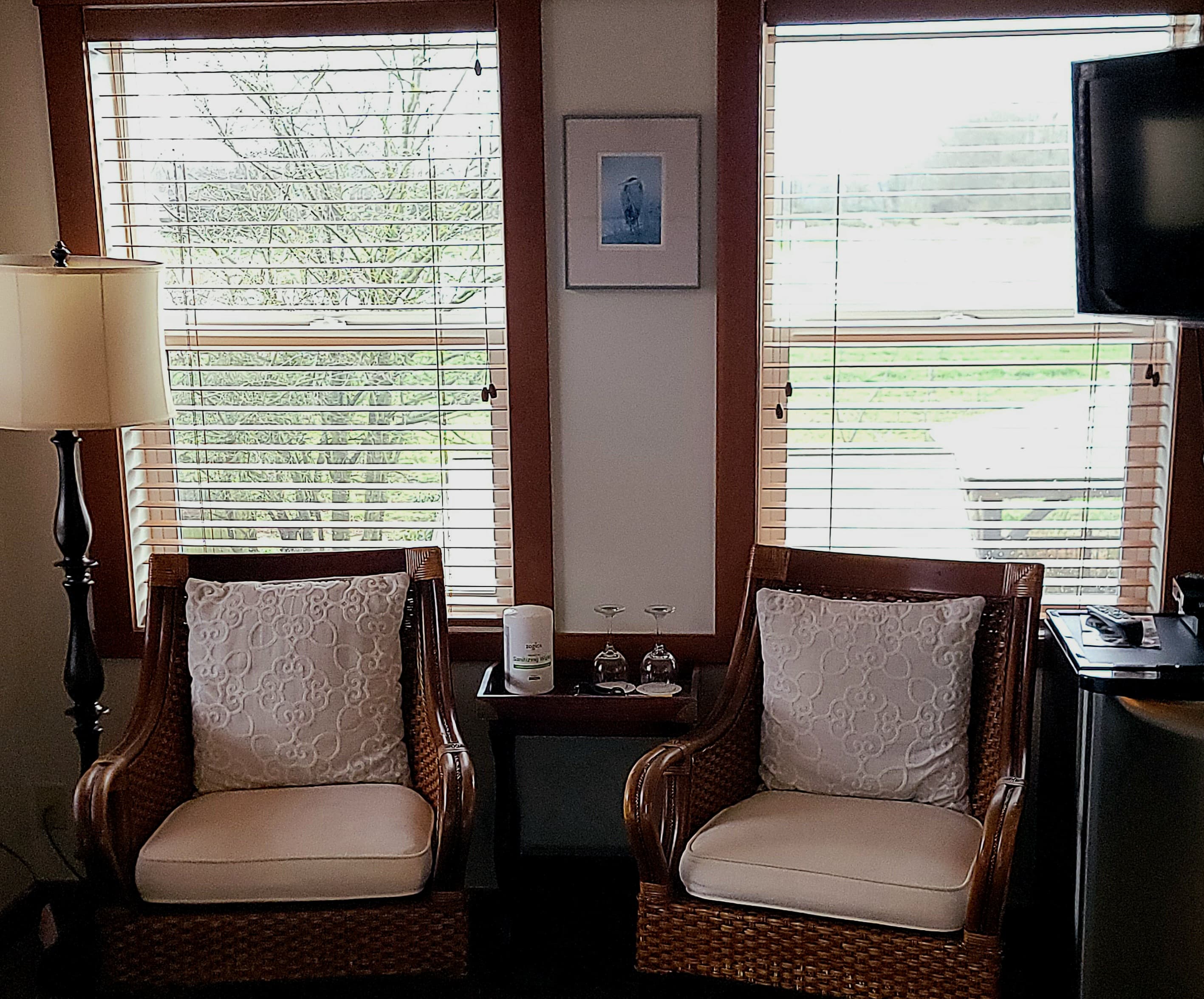 A cozy seating area with two wicker chairs, a side table, a lamp, and windows with blinds letting in natural light.