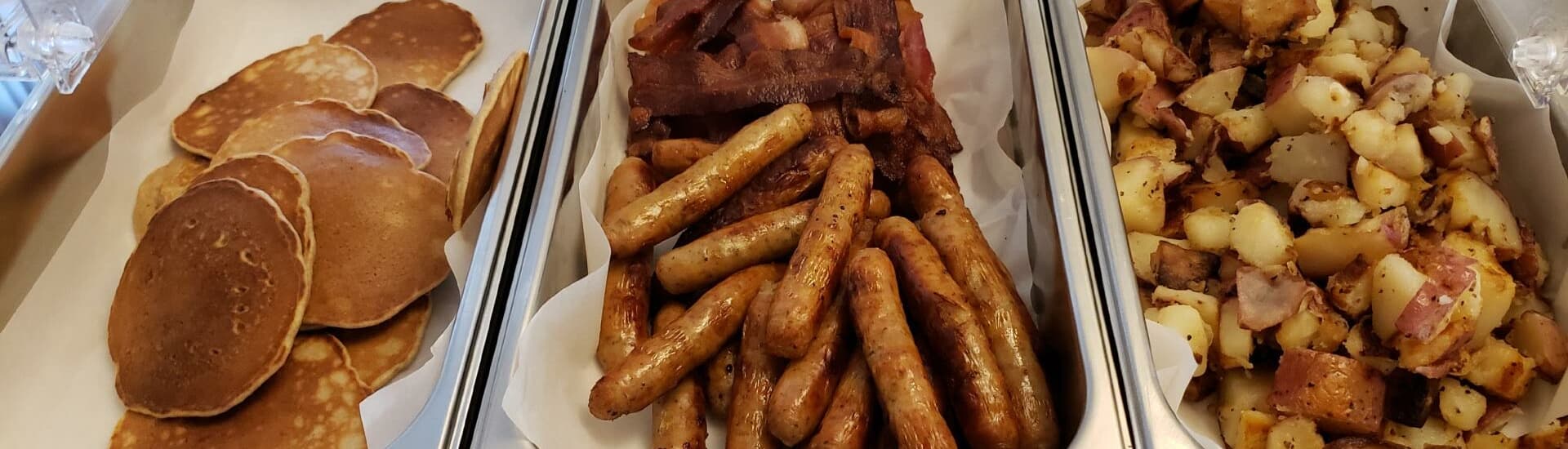 A selection of pancakes, sausage links, bacon, and diced potatoes in serving trays.