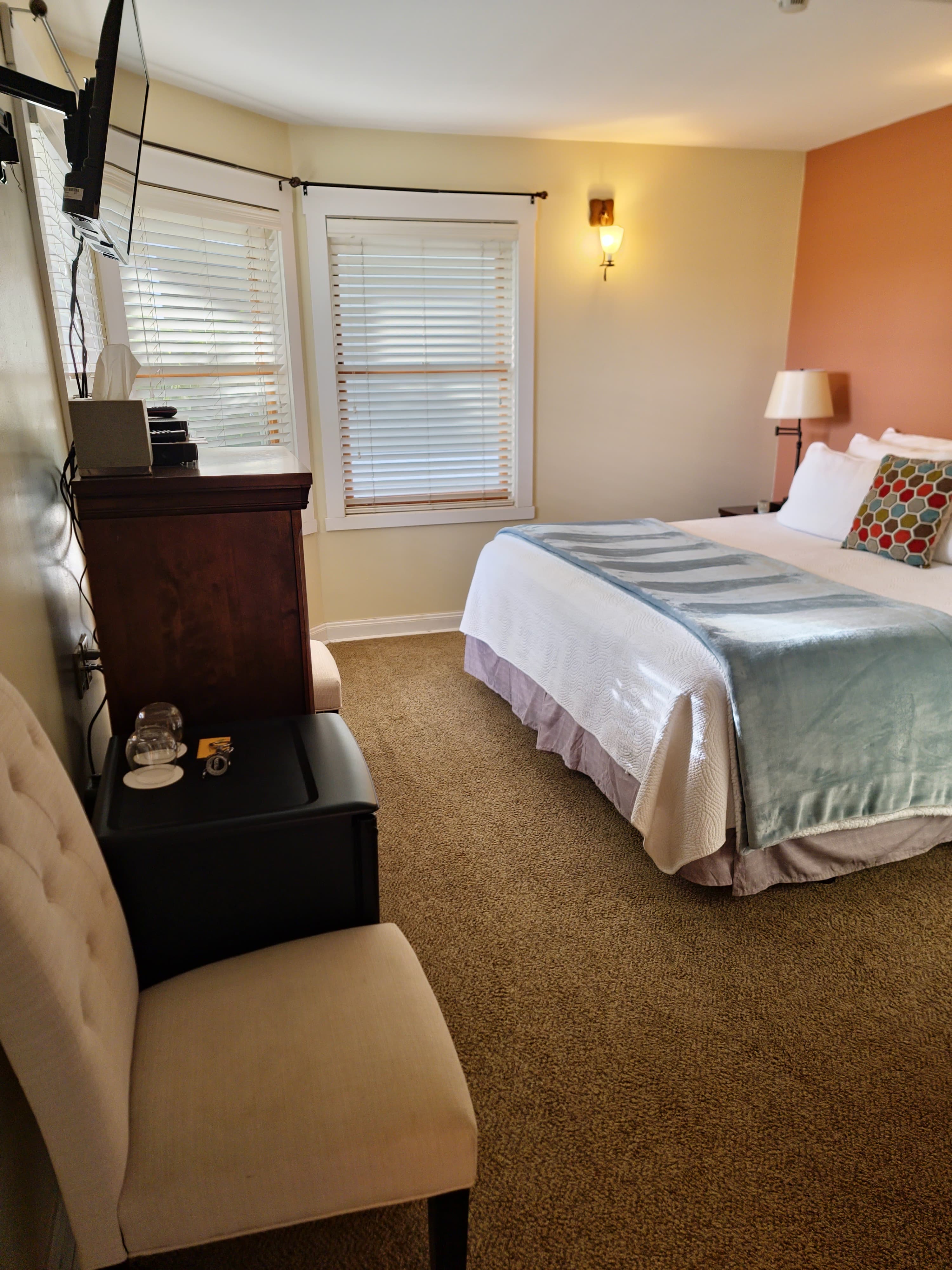 A cozy bedroom with a bed, a chair, a small nightstand, and windows with blinds.