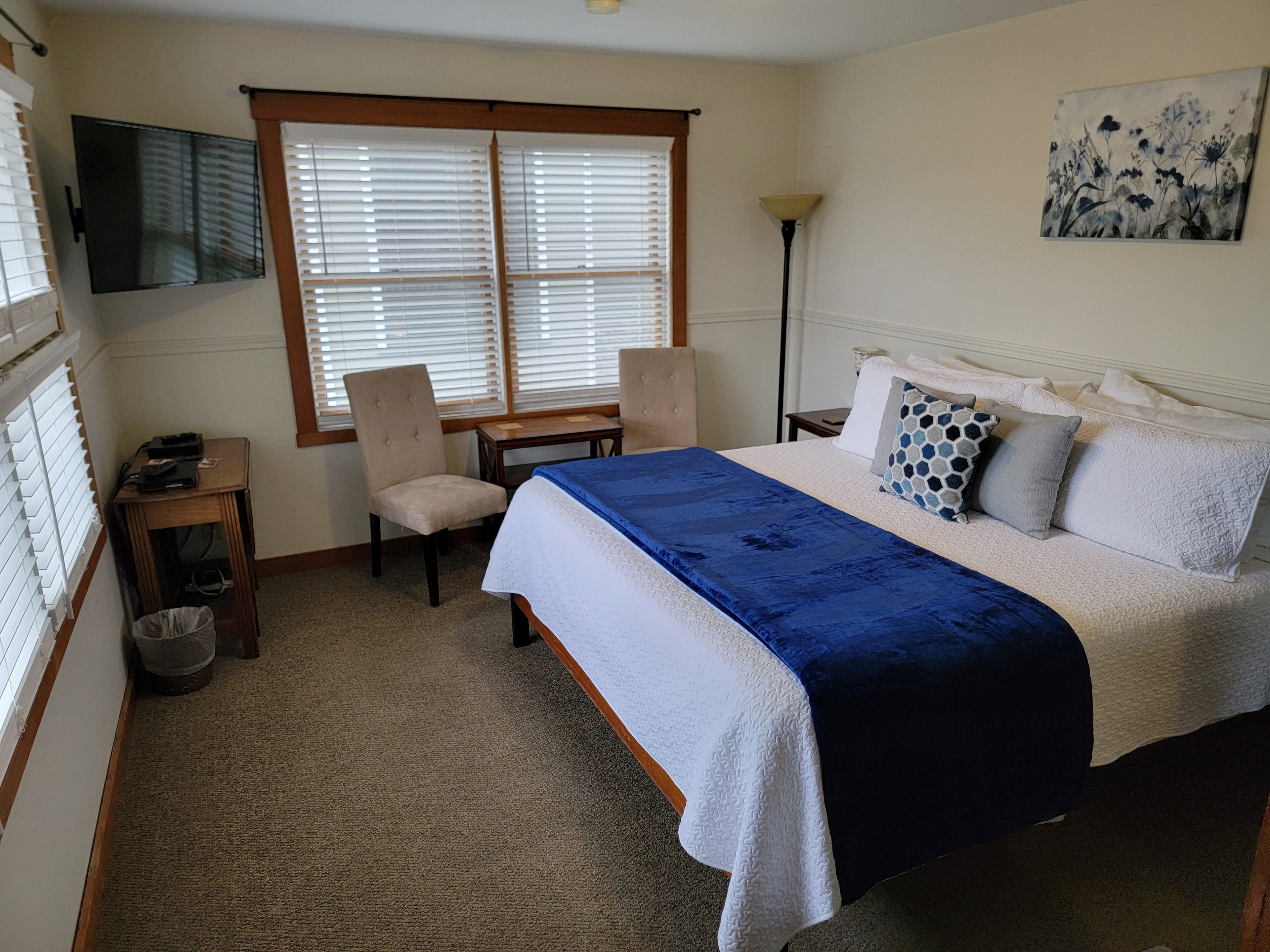 A cozy bedroom featuring a bed with a blue throw blanket, two chairs, and a TV mounted on the wall, with natural light from the windows.