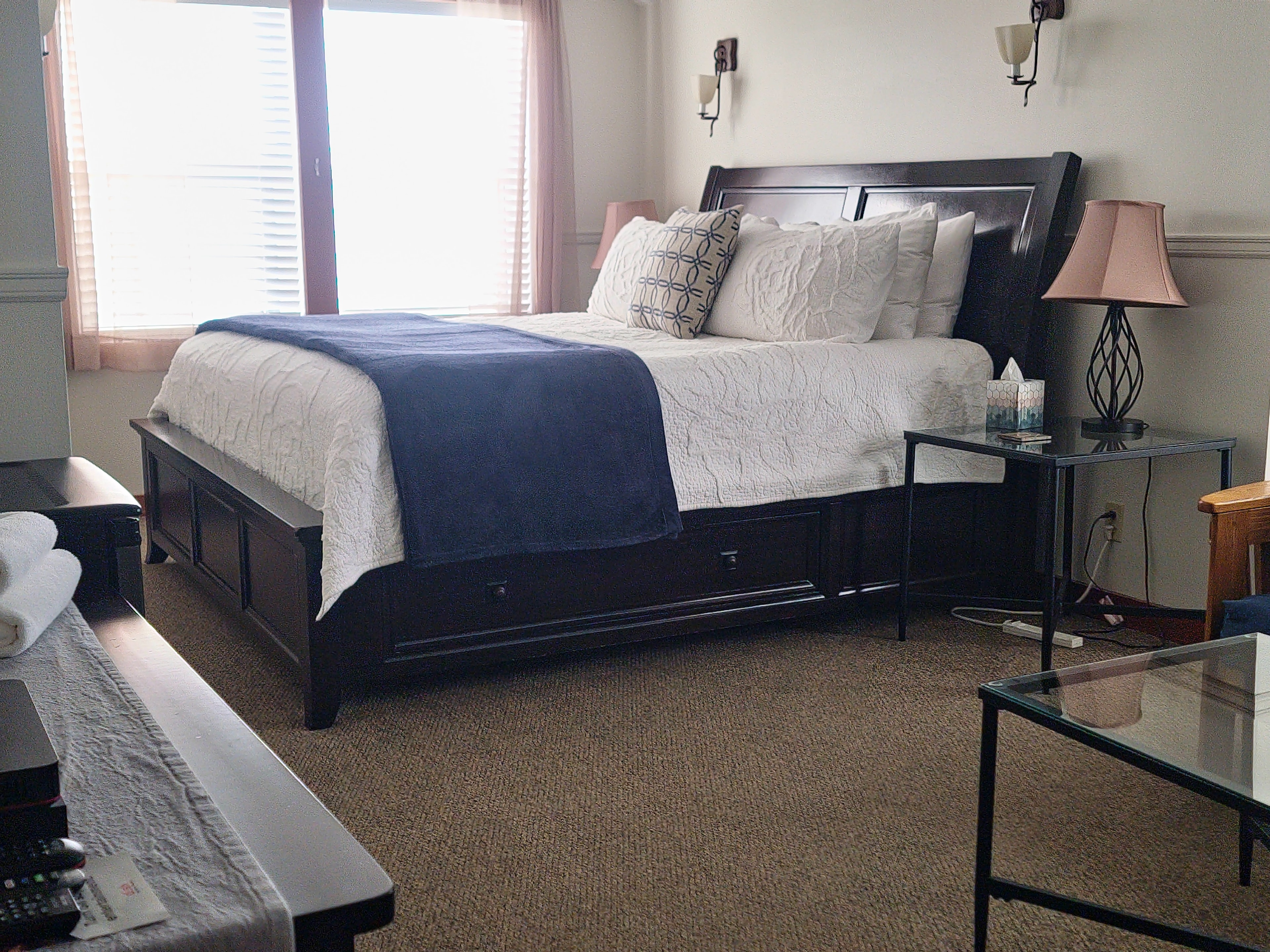 A cozy bedroom featuring a white bedspread, decorative pillows, a nightstand, and natural light from a window.