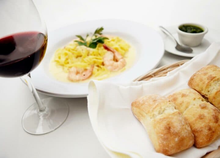 A glass of red wine accompanied by shrimp fettuccine and fresh bread rolls.