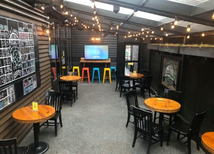 Indoor and outdoor seating area with wooden tables, colorful stools, a chalkboard menu, and string lights.