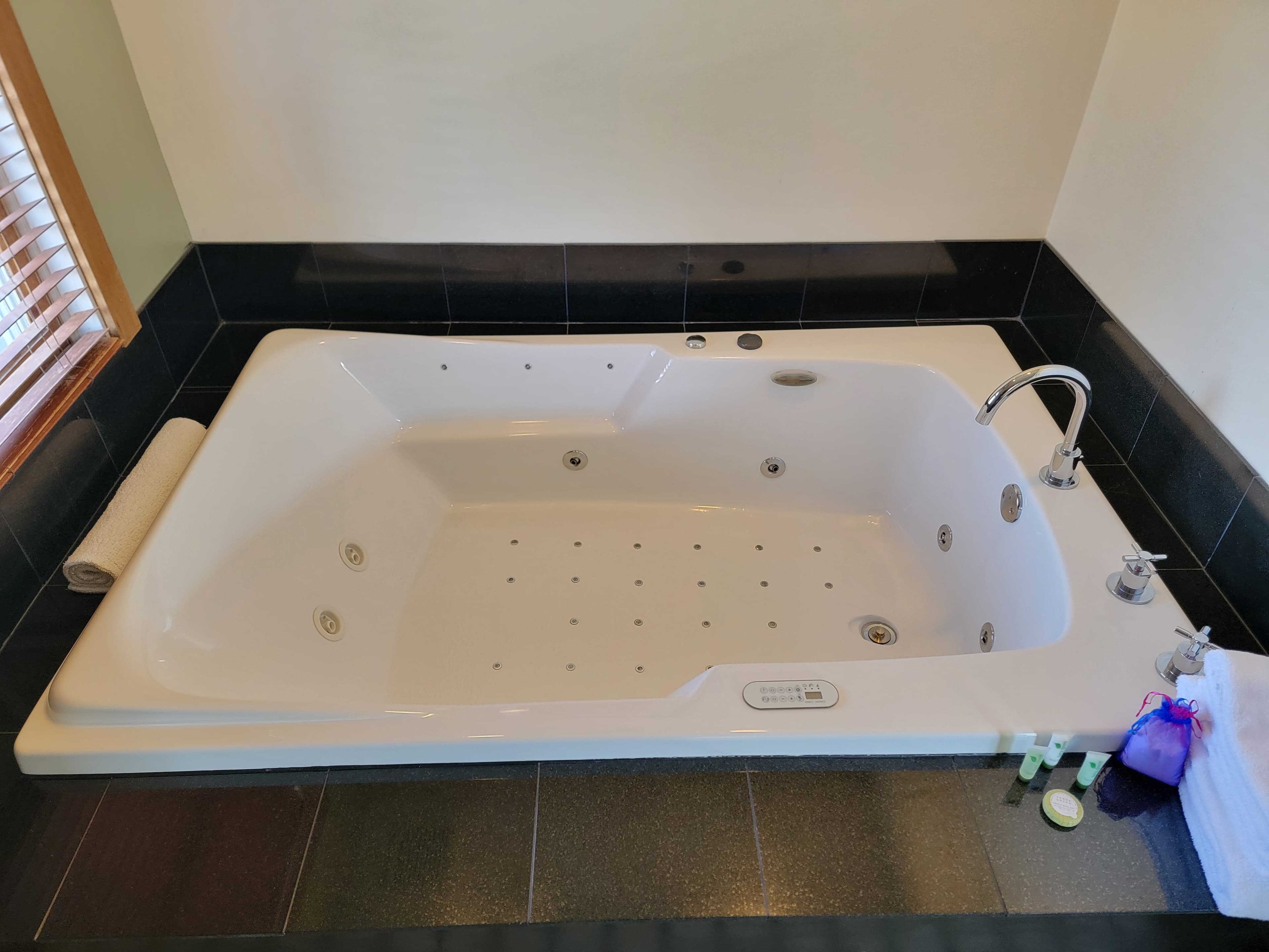 A white jacuzzi tub with a chrome faucet on a dark tiled surface, next to towels and toiletries.
