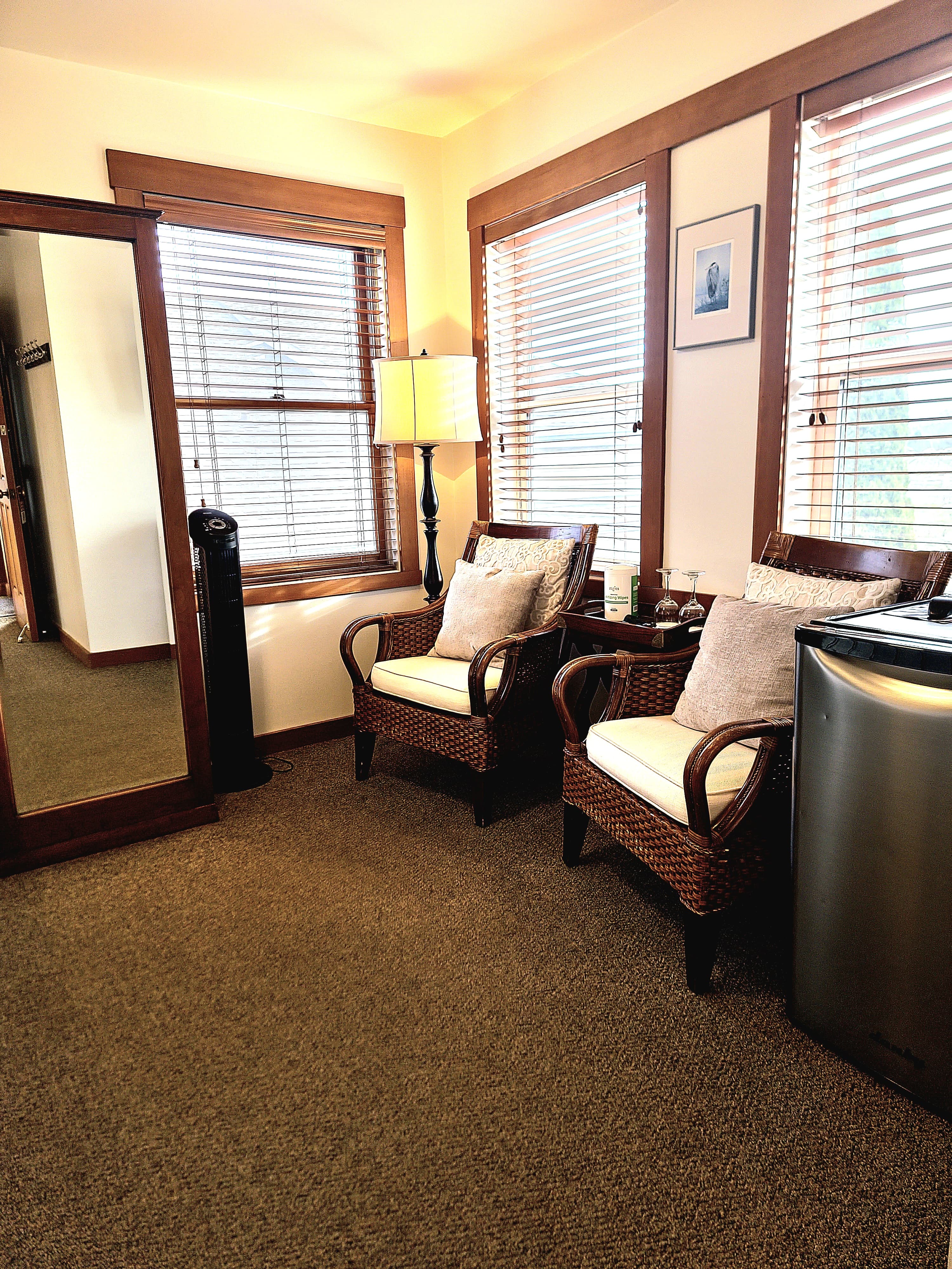 A cozy sitting area with two wicker chairs, a lamp, and a mirror, illuminated by natural light from the windows.