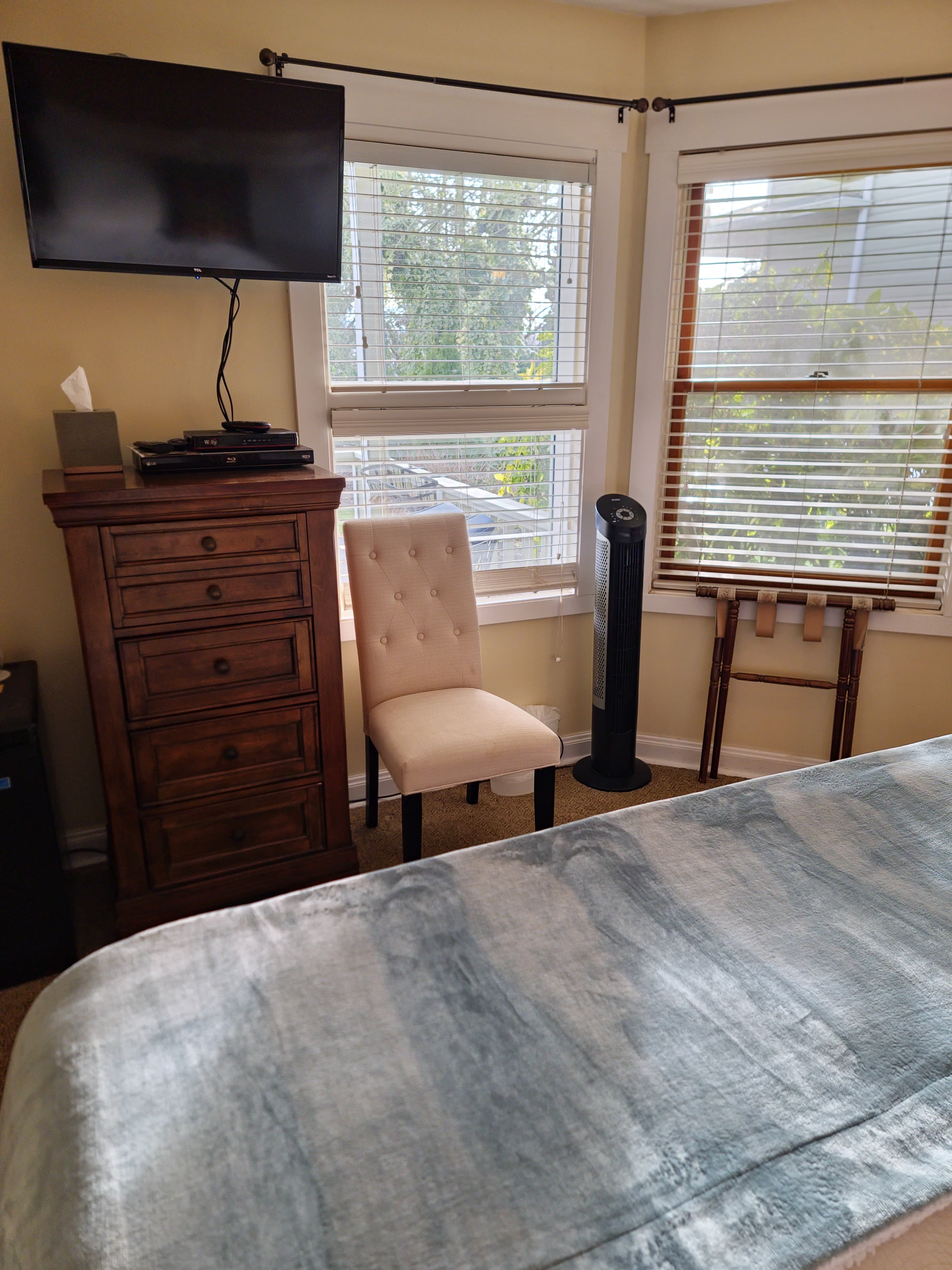 A cozy bedroom featuring a wooden dresser, a chair, a TV mounted on the wall, and windows with blinds.