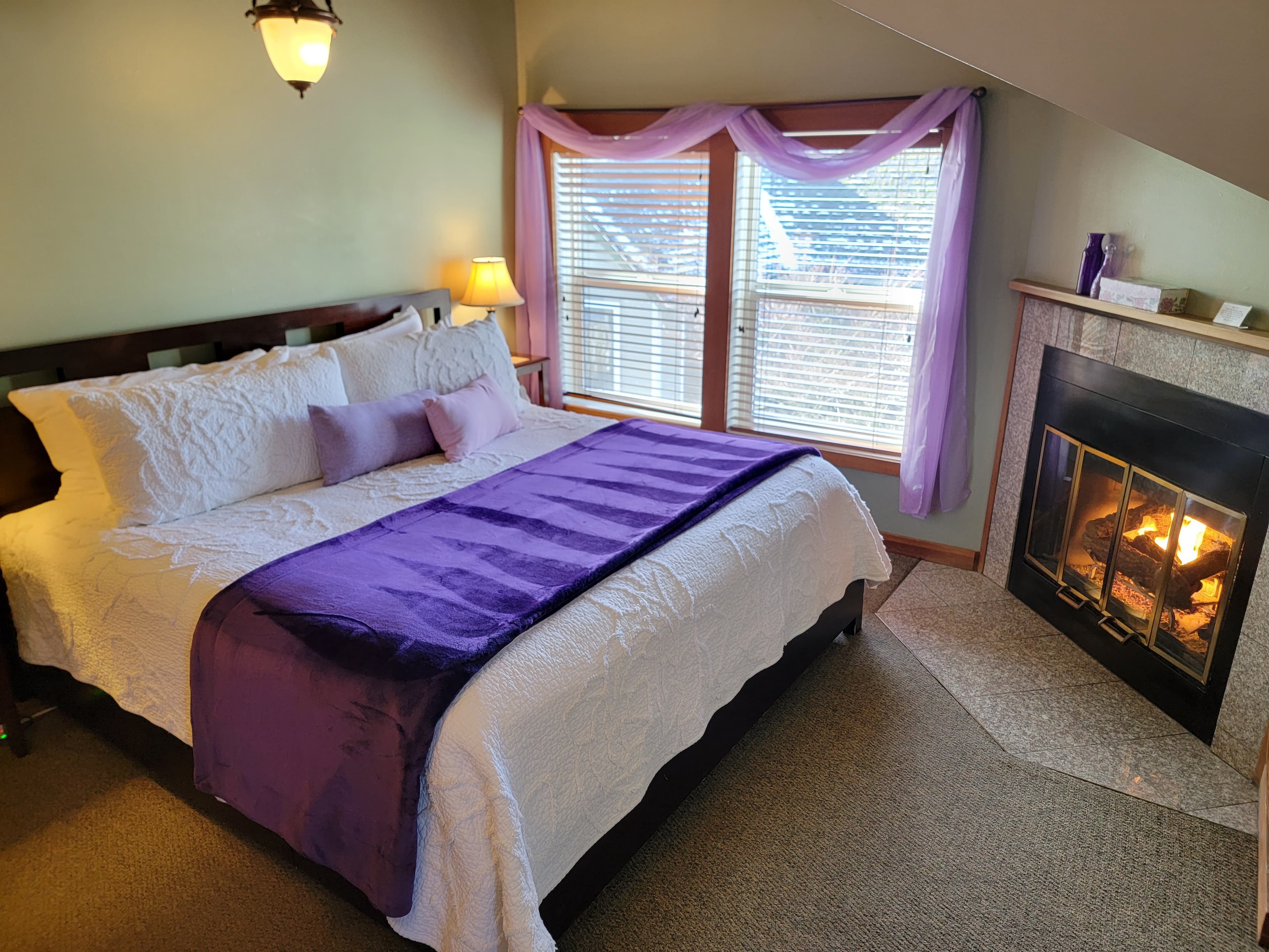 A cozy bedroom with a bed adorned in white and purple, featuring a fireplace beside large windows.