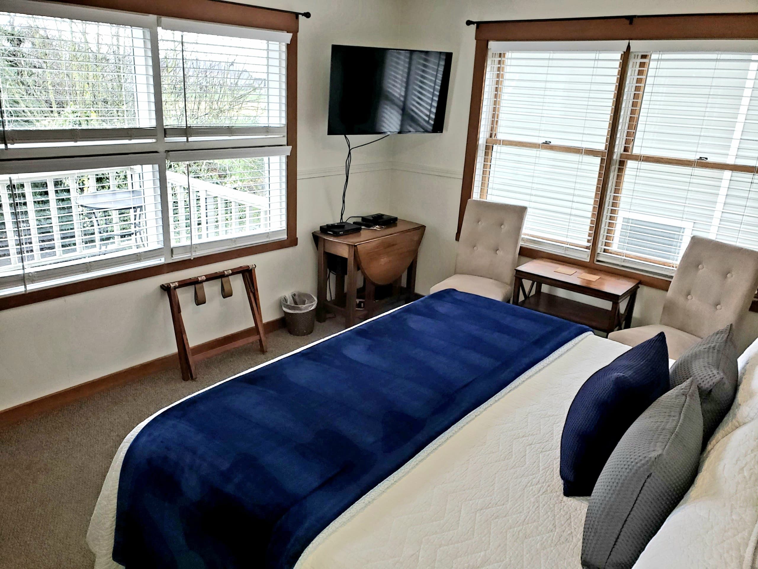 A cozy bedroom featuring a bed with a blue blanket, two chairs, a television, and large windows with natural light.