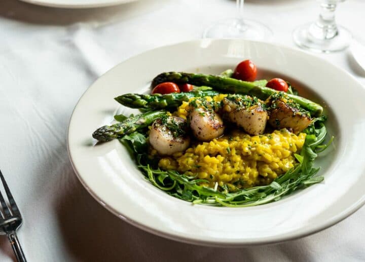 A plate of saffron risotto topped with grilled scallops, asparagus, and cherry tomatoes on a bed of arugula.