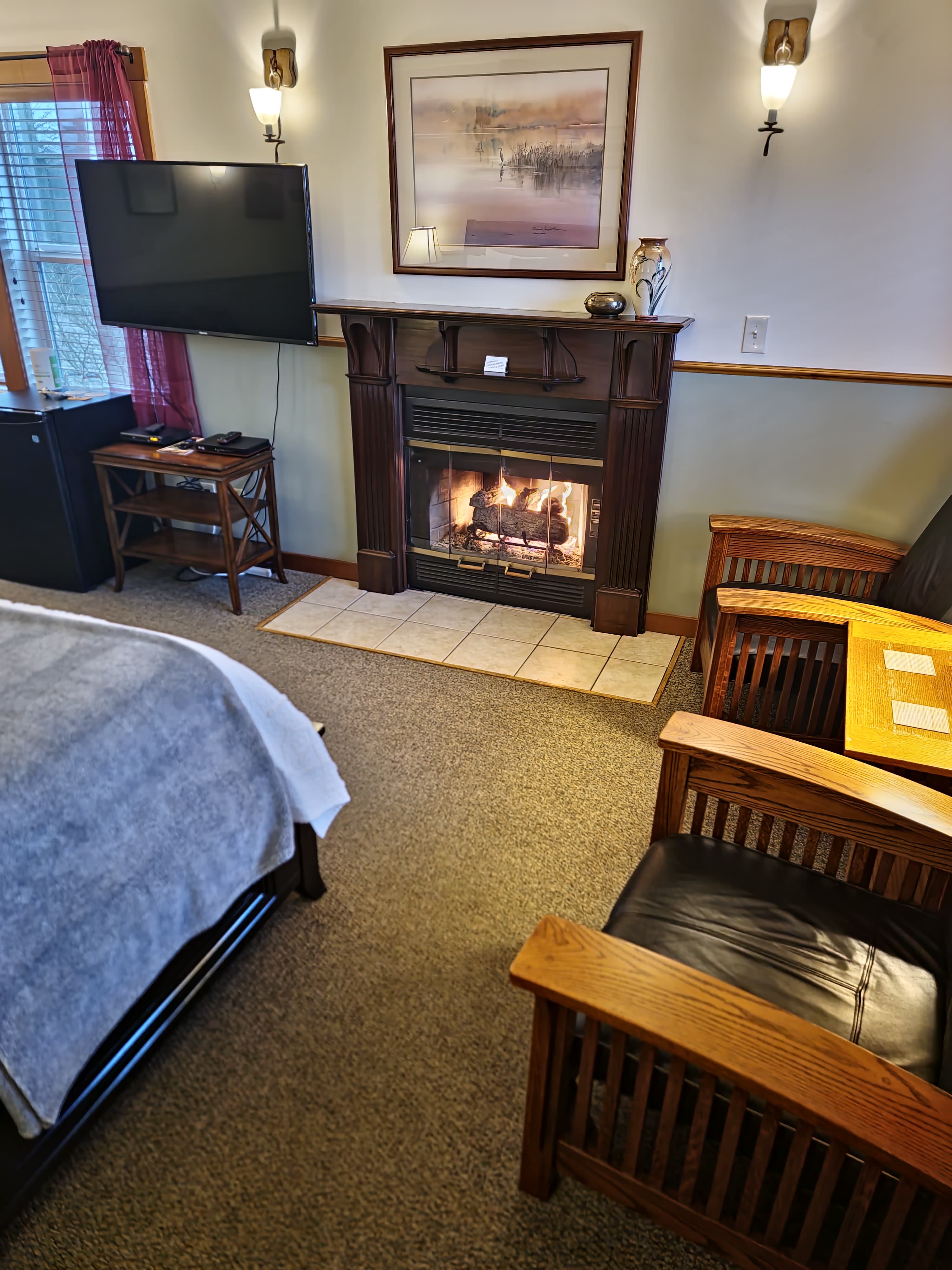 A cozy room featuring a fireplace, two armchairs, a TV, and a bed with a soft blanket.